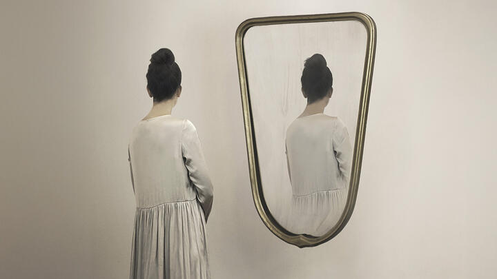 A woman looking at a half length mirror only to see her reflection facing away from her 